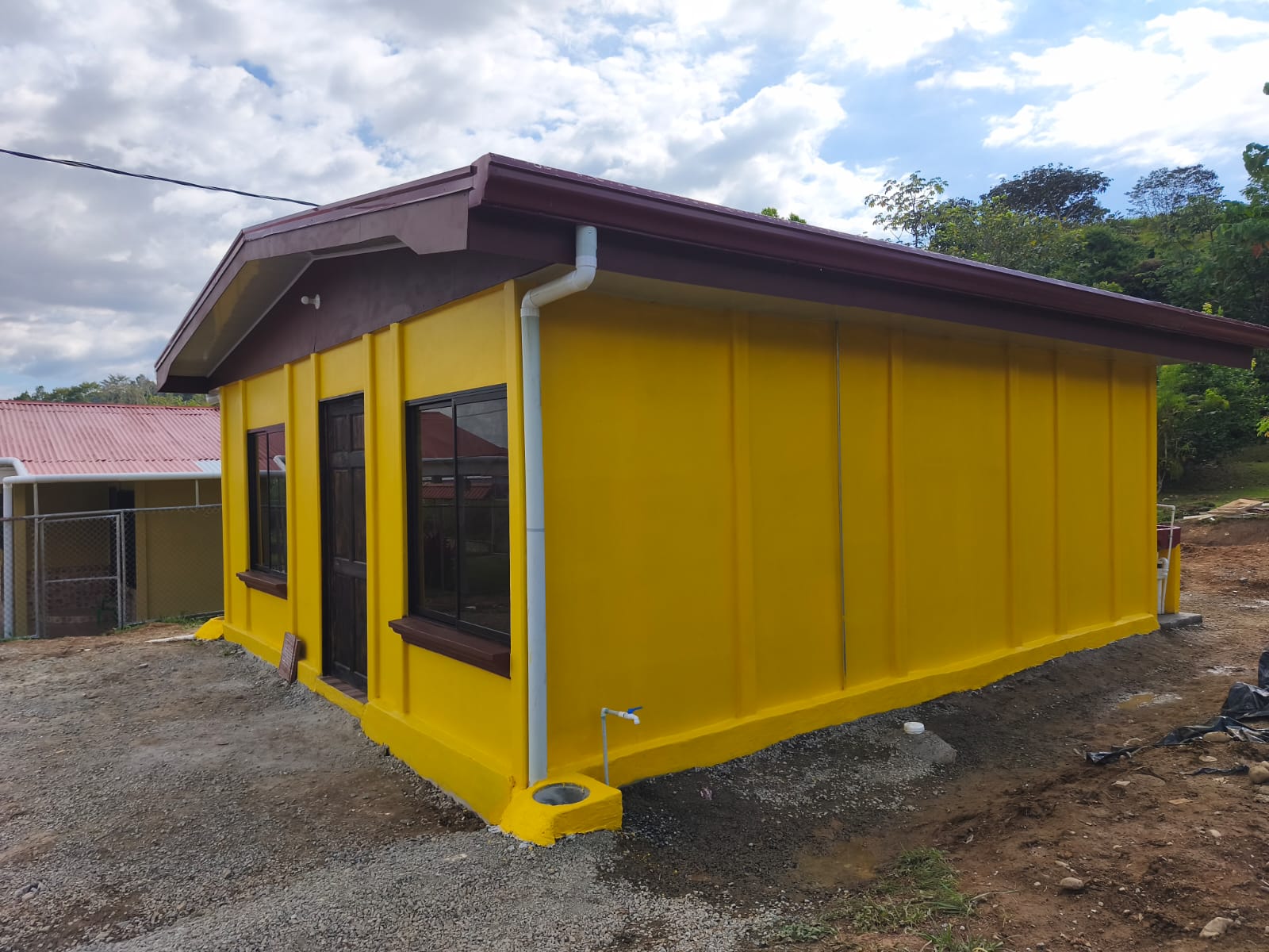 Vista frontal de casa de bono de vivienda prefabricada color amarillo intenso con techo marrón oscuro y ventanas de aluminio negro